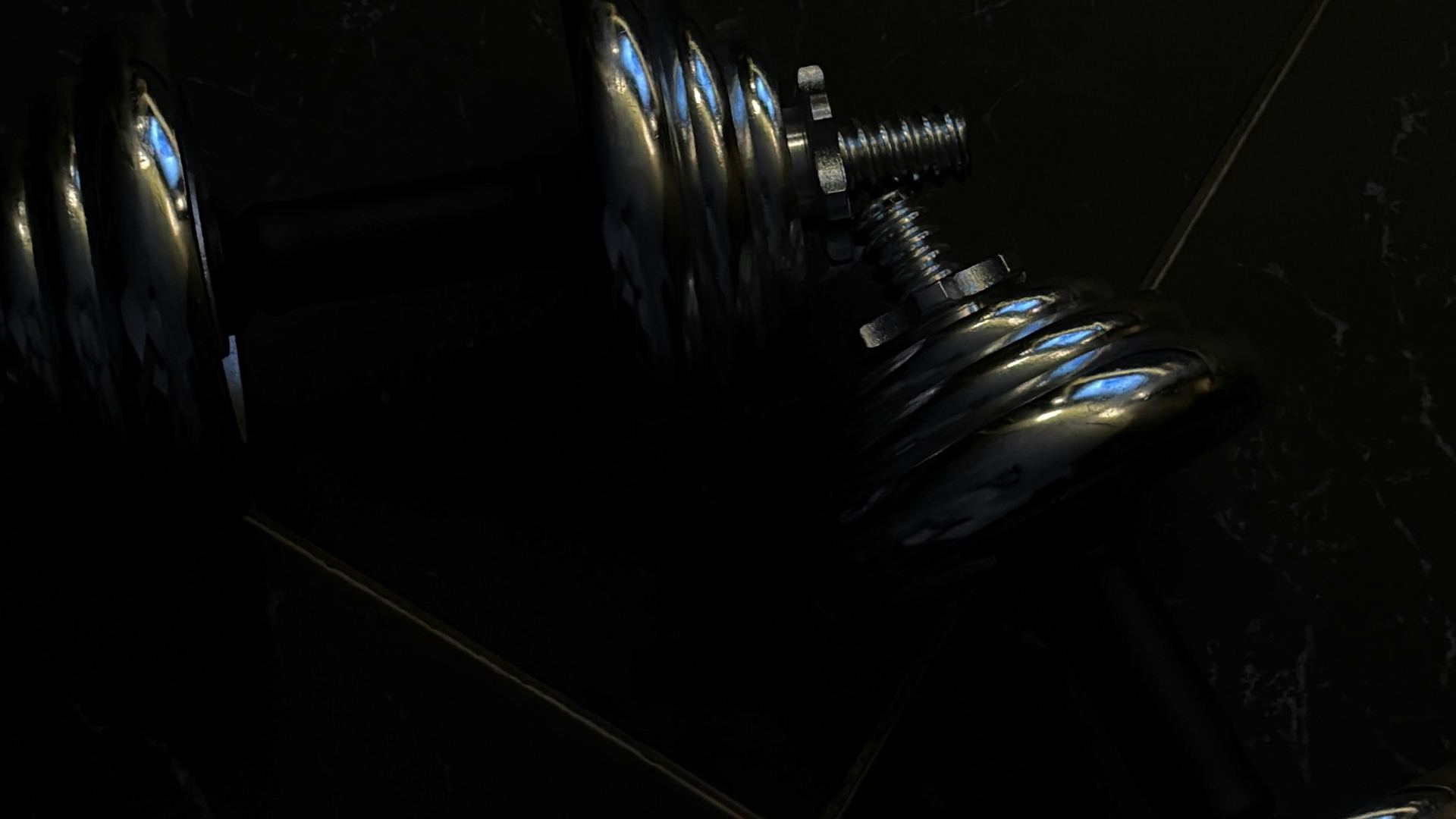 Close-up of chrome dumbbells resting on a dark tiled floor in low light